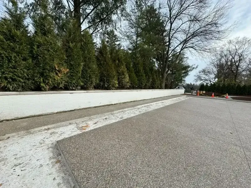 A concrete road with a long white wall and evergreen trees. Orange cones are visible in the distance.