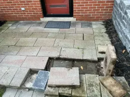 Damaged brick patio in front of a brick building with a red door. Several paving bricks are missing or displaced.