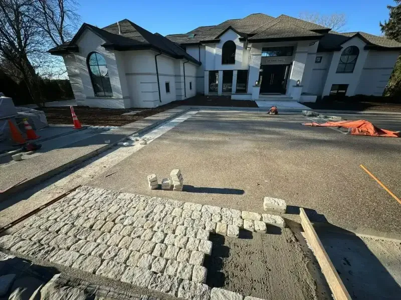 Driveway construction in front of a large white house. A partially finished cobblestone walkway and driveway are visible.
