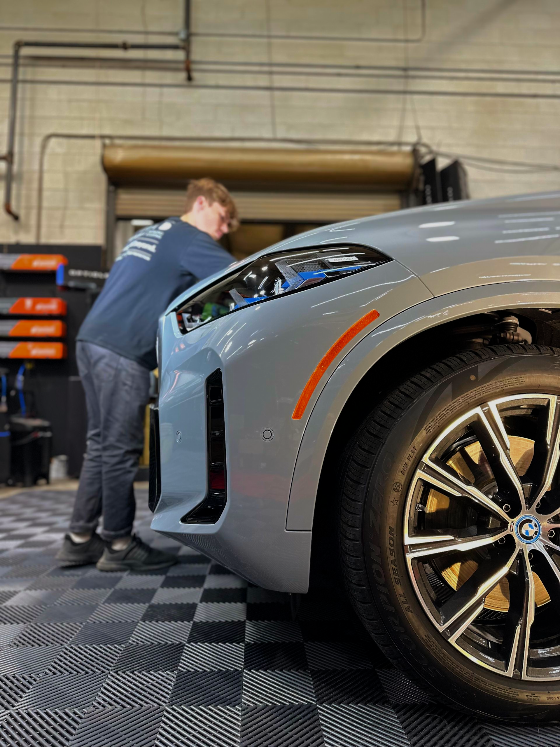 Man working on the front of a gray BMW in a garage; the car's headlight is illuminated.