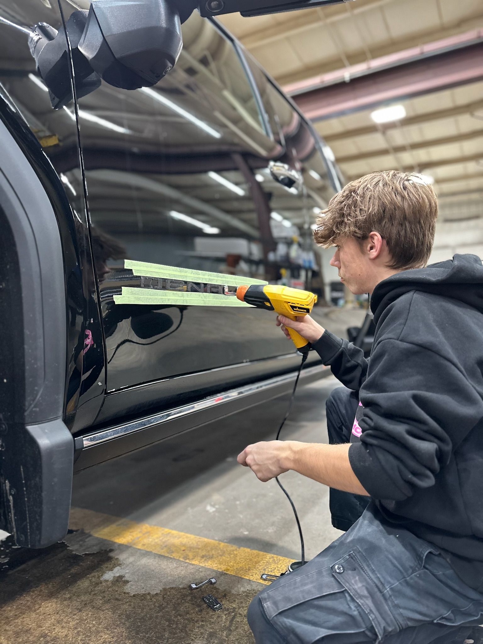 Young person uses a heat gun on a black car door in a shop.