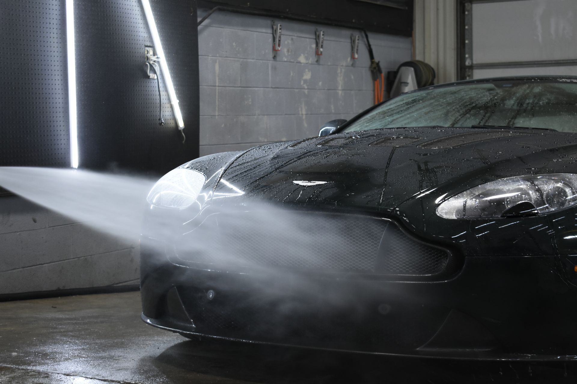A black sports car being washed with a high-pressure hose in a garage, water spraying from the nozzle.