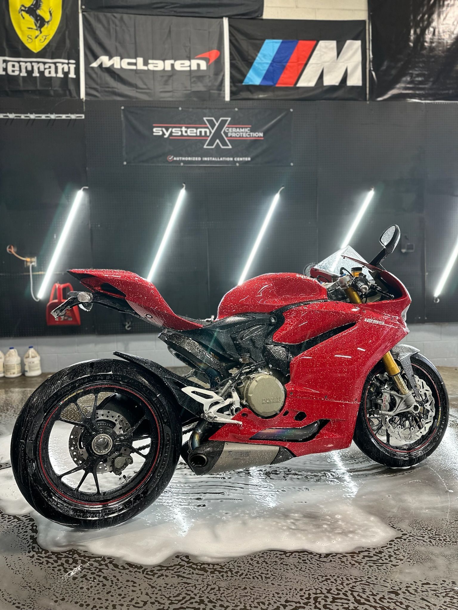 Red Ducati motorcycle being washed with foamy soap. Garage setting with flags in background.