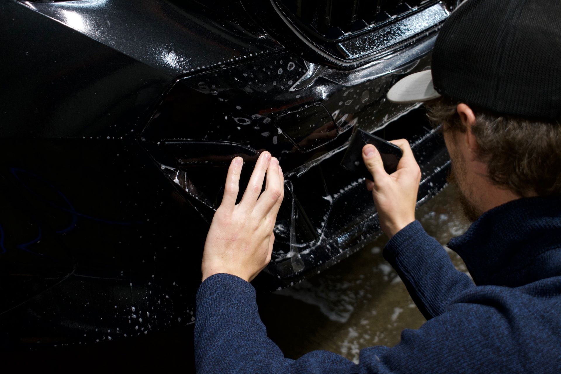 Person applying clear protective film to black car's front bumper.