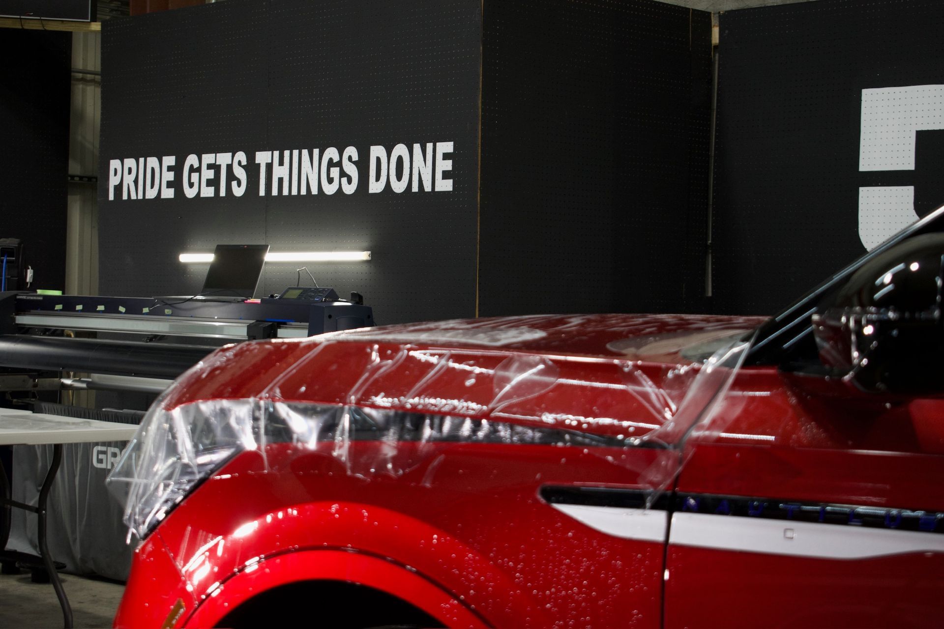 Red car with clear protective film being applied; workroom, sign reads 