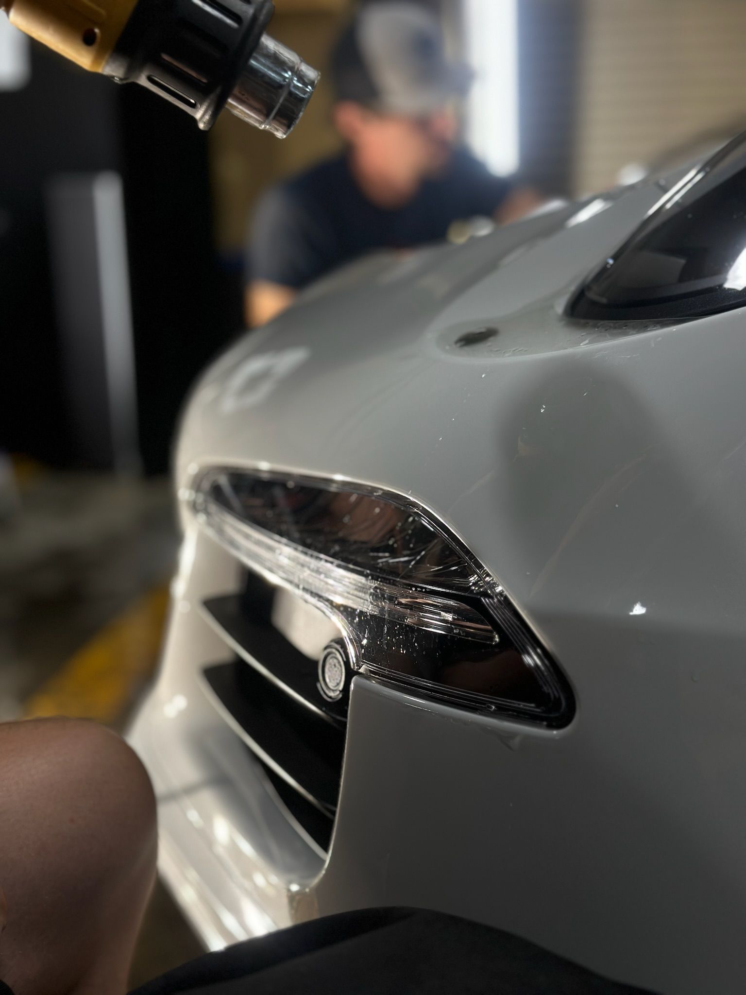 Person using a heat gun on a car's headlight, possibly applying film. White car in a garage.
