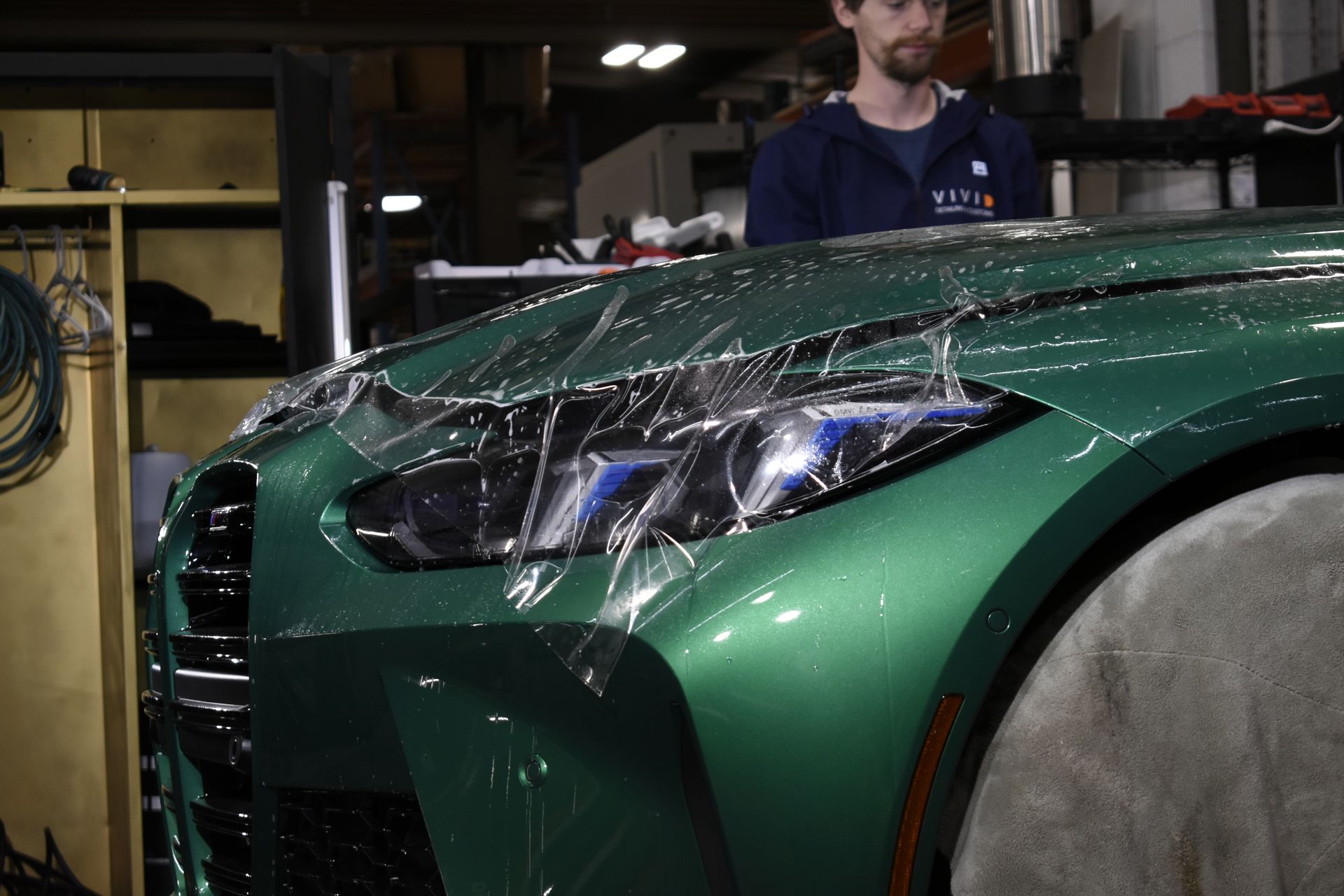 Green sports car with protective film being applied, man in background.