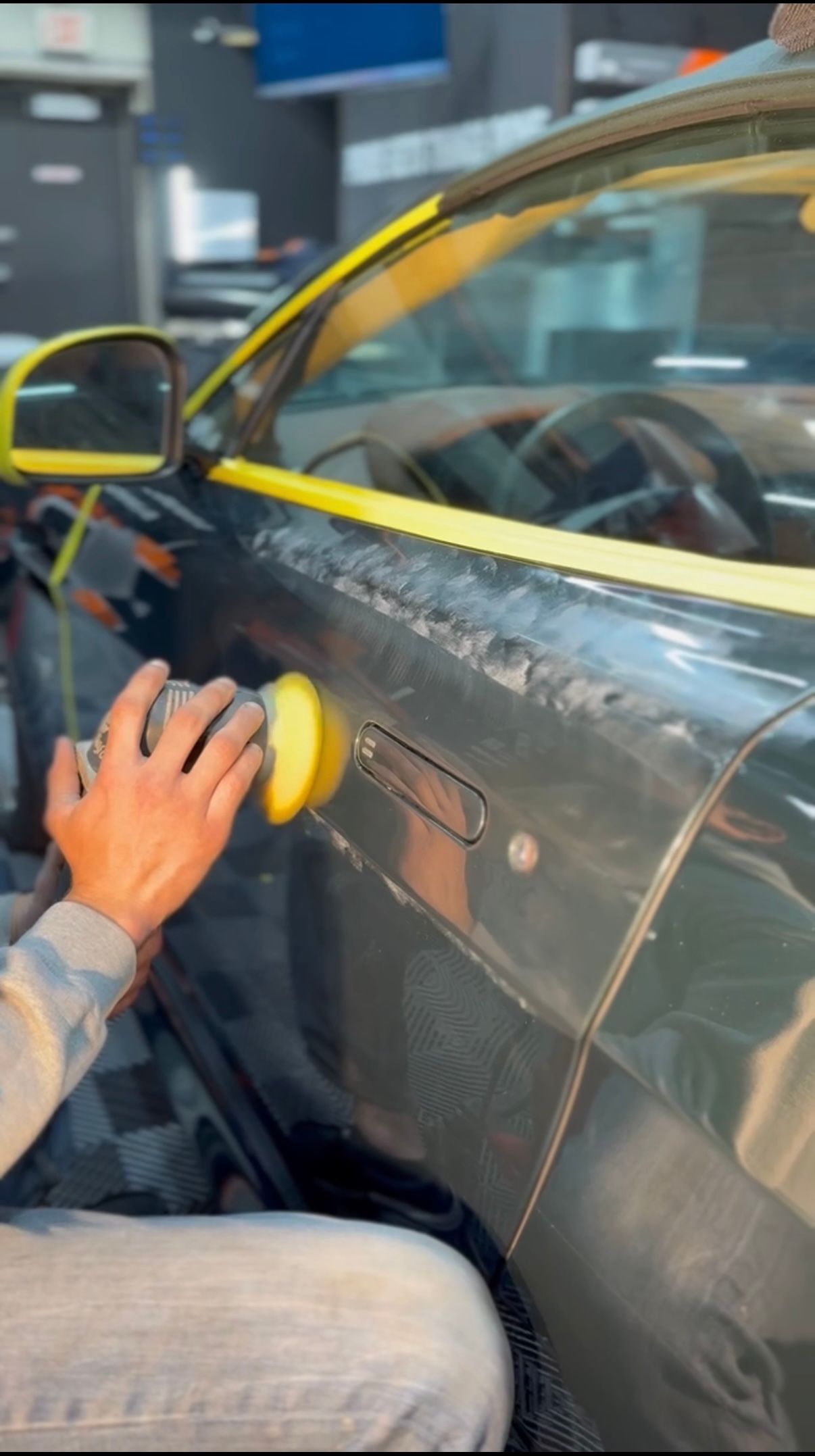 Person polishing a black car door with a yellow buffer in a garage.