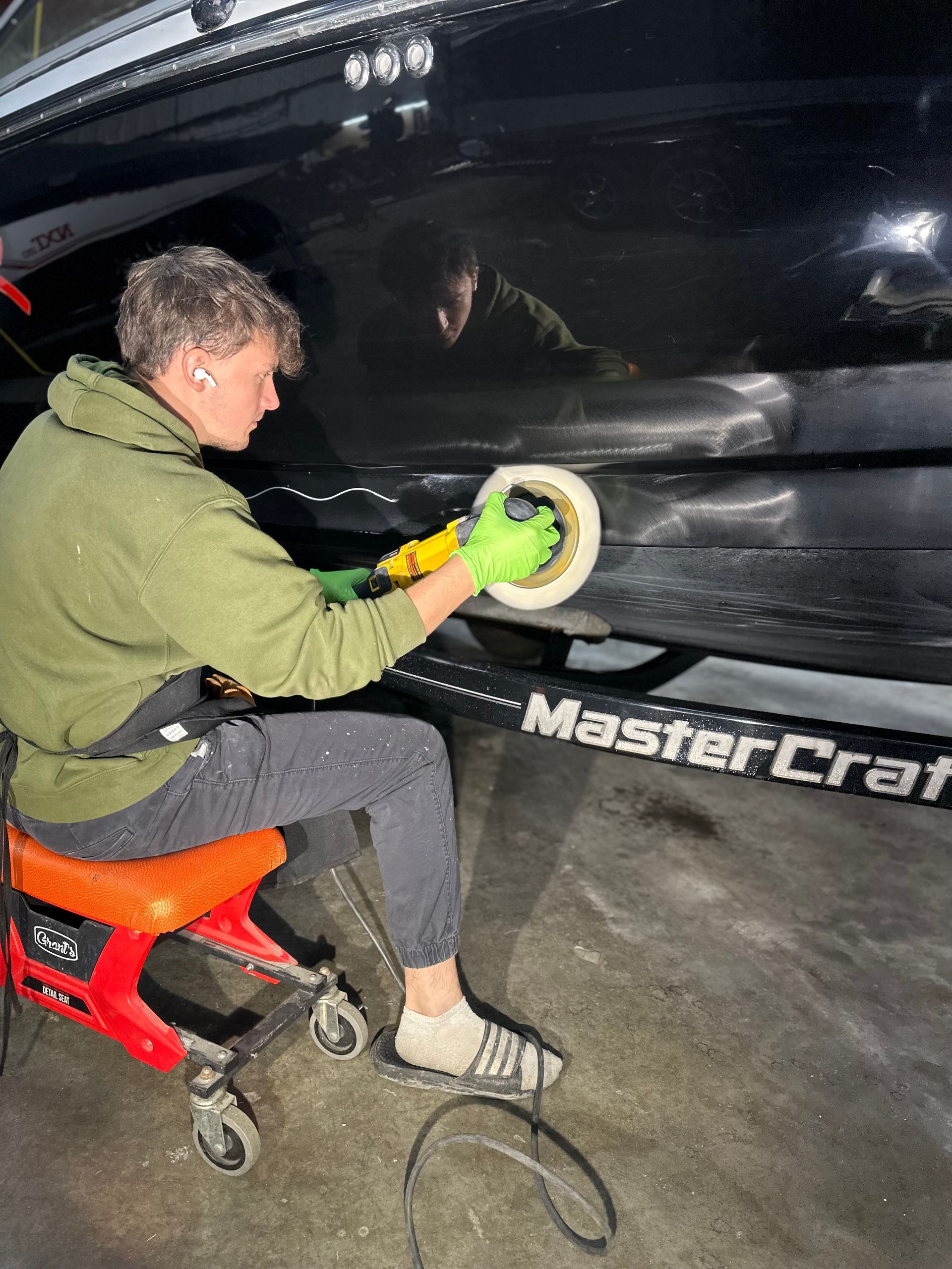 Person polishing black boat with a rotary buffer. They're wearing green hoodie, on a rolling stool, indoors.