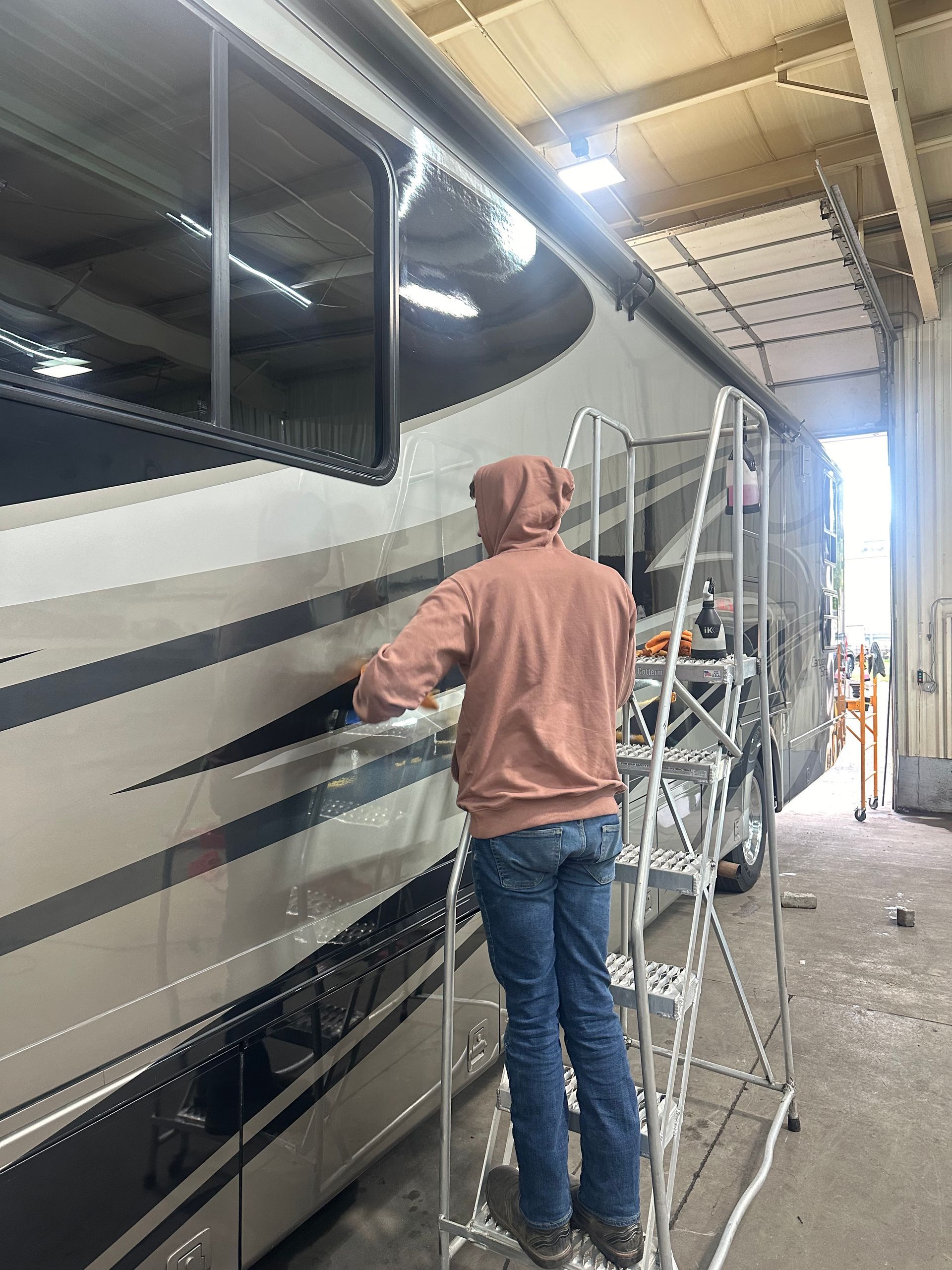 Person on a ladder working on the side of a large beige and black RV inside a garage.