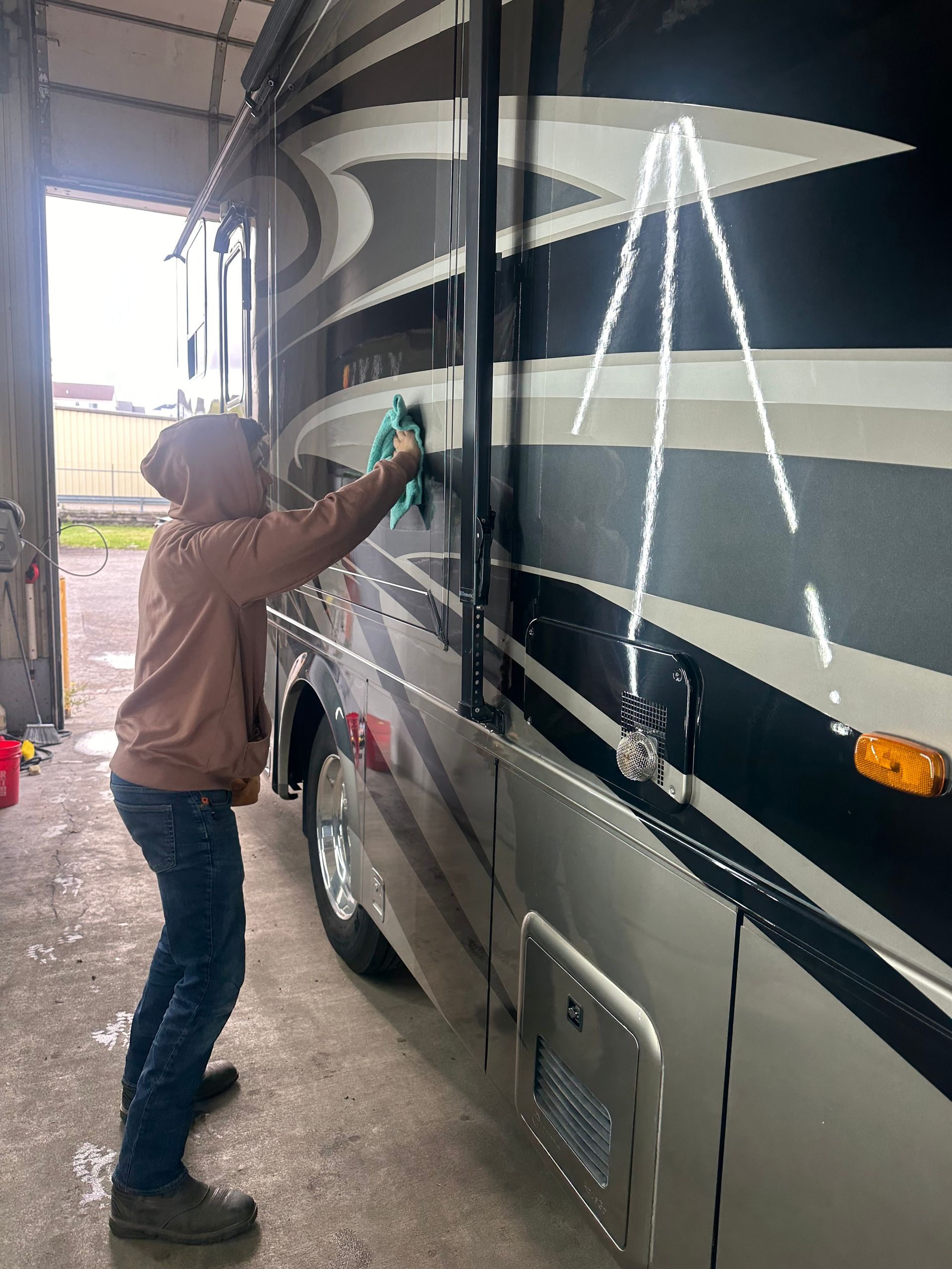 Person washing the side of a large RV in an enclosed bay; silver, black, and beige colors.