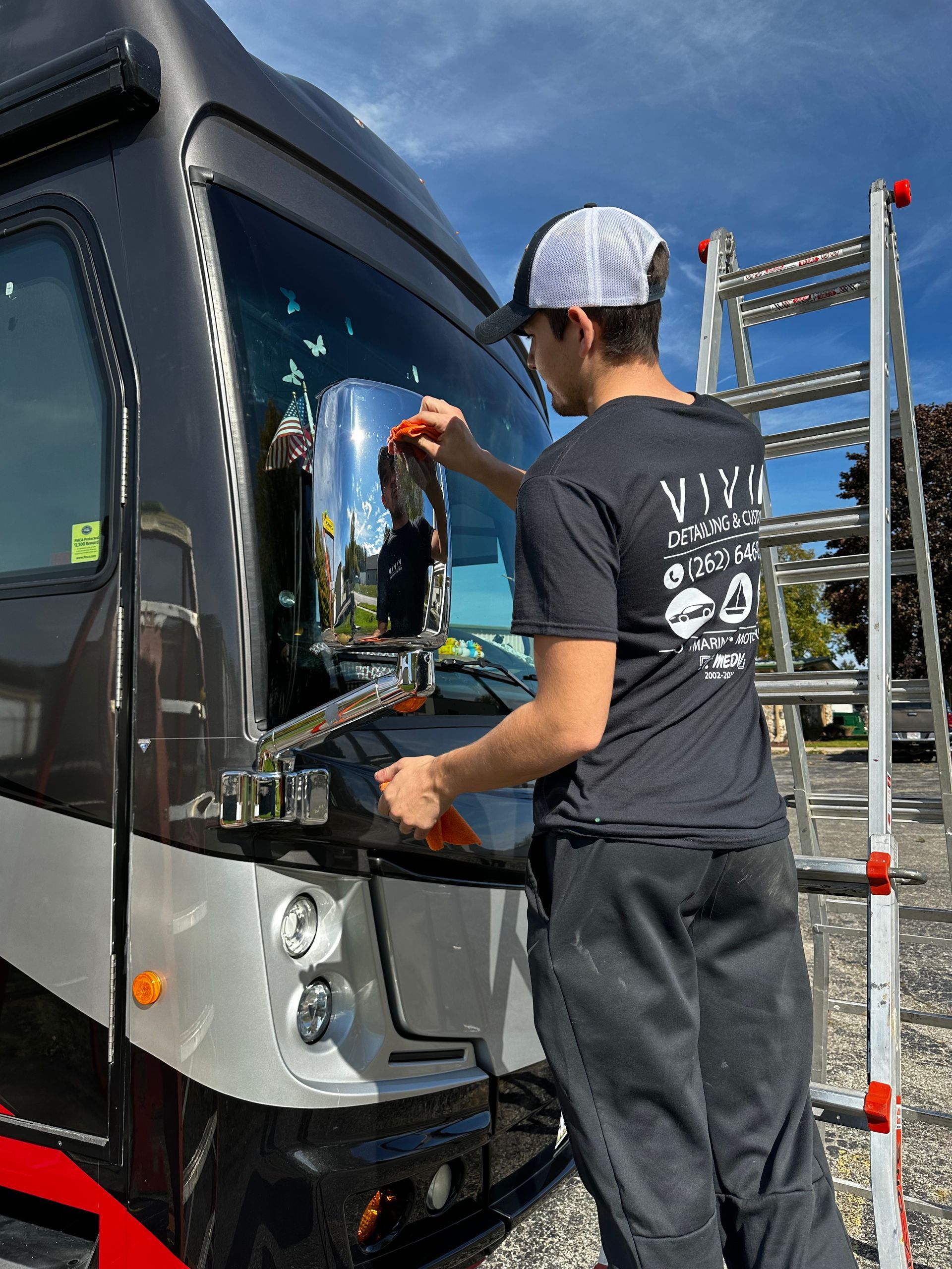Man cleaning RV windshield with ladder on a sunny day.