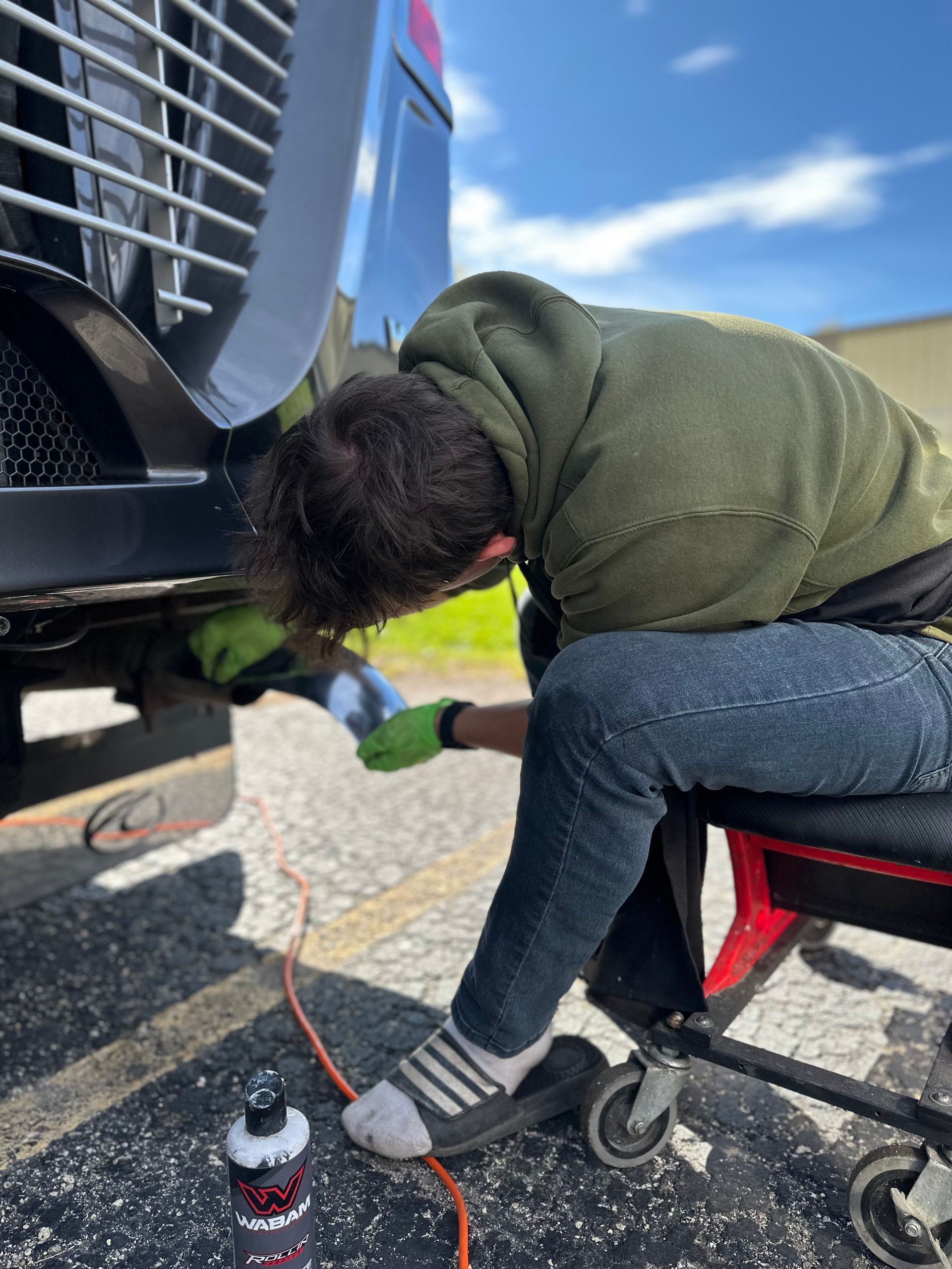 Man washing undercarriage of a black vehicle, seated on a rolling stool, wearing green gloves.
