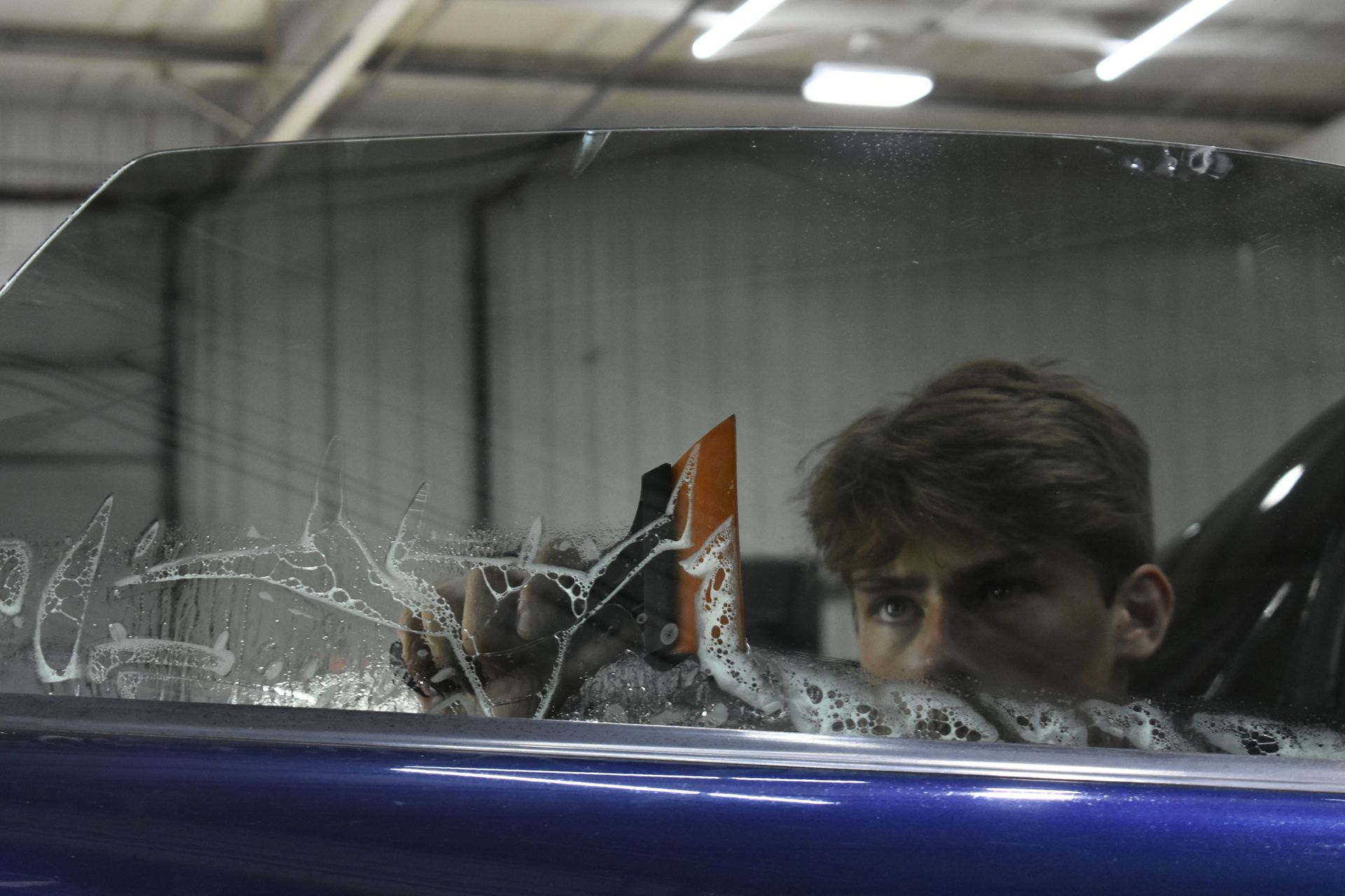 Man applying window tint to a car inside a garage. He's looking at the glass and holding a squeegee.