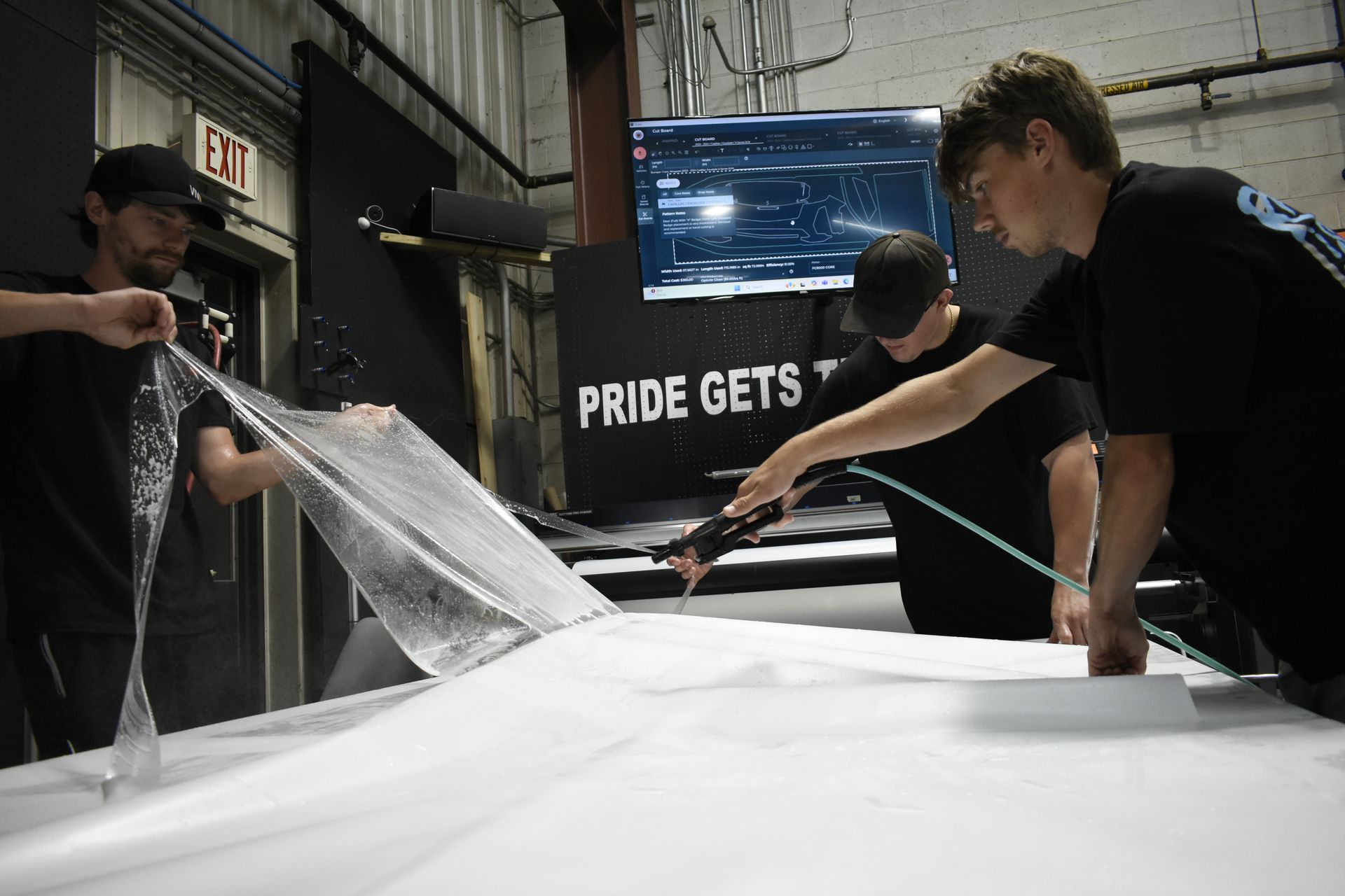 Three men applying large white film to a car in a shop, using tools and a computer monitor.