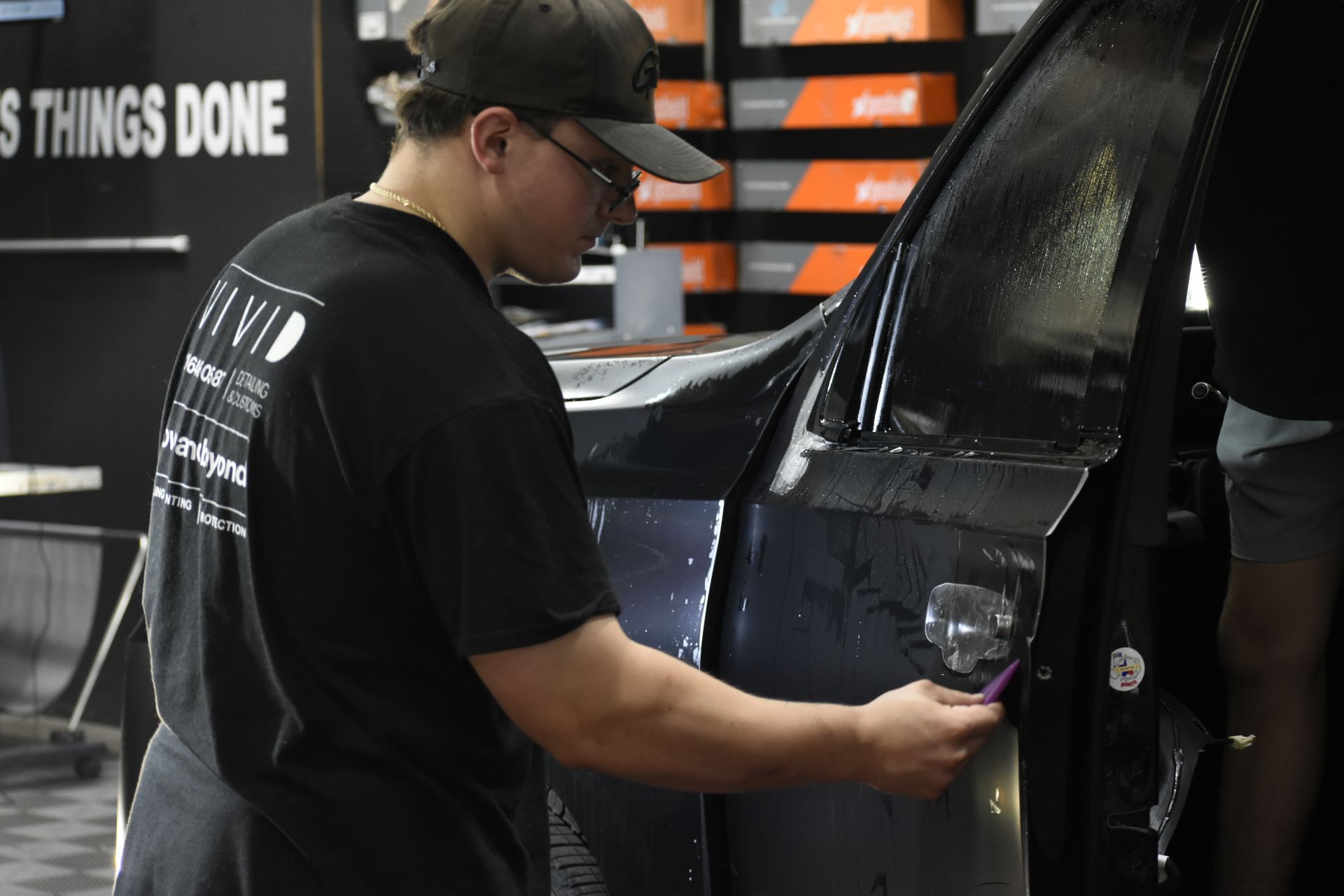 A man in a black shirt and cap applying film to a car door, indoors.