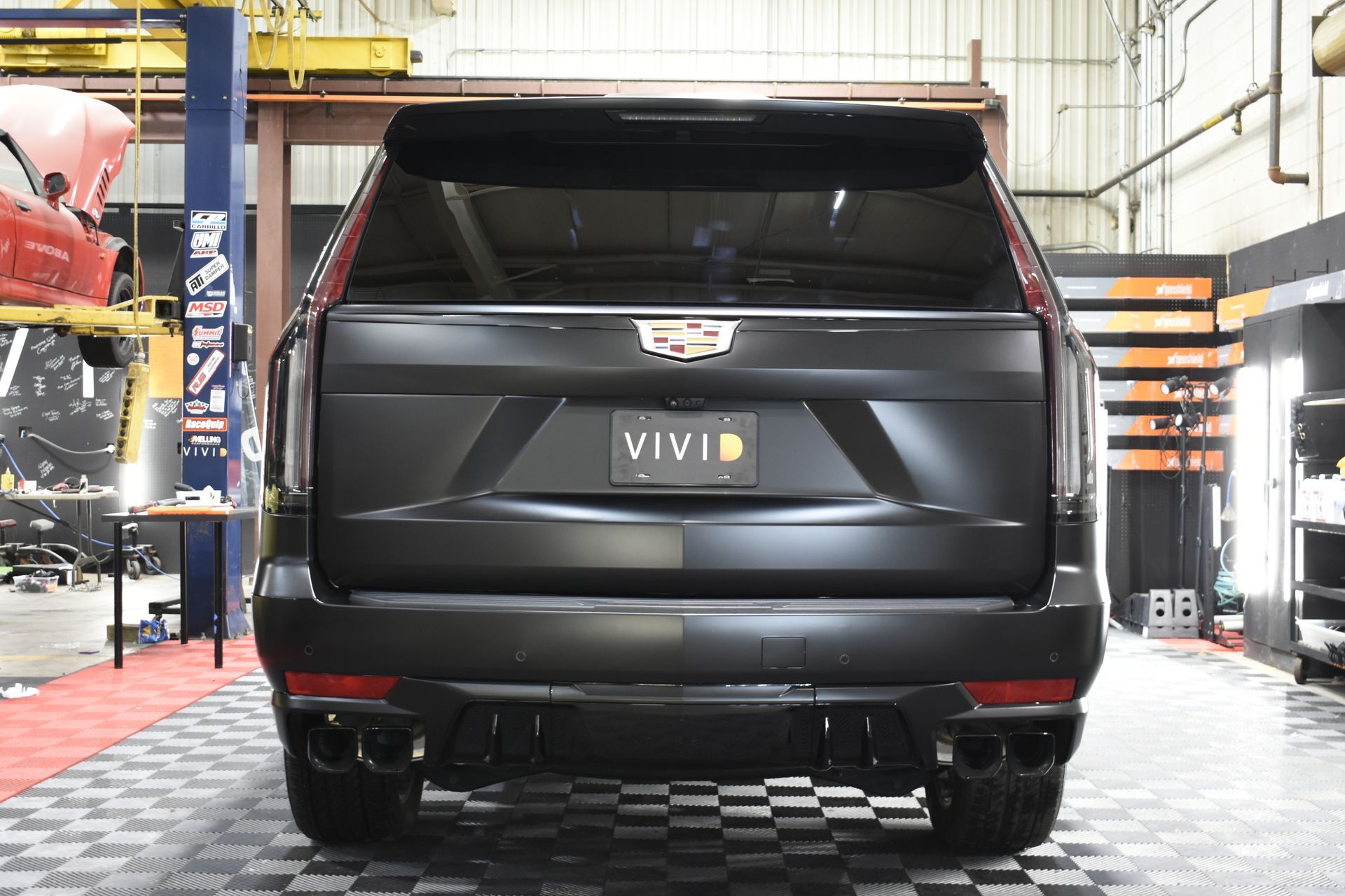 Black Cadillac Escalade rear view in a garage. Matte black finish, dual exhaust, vivid license plate.