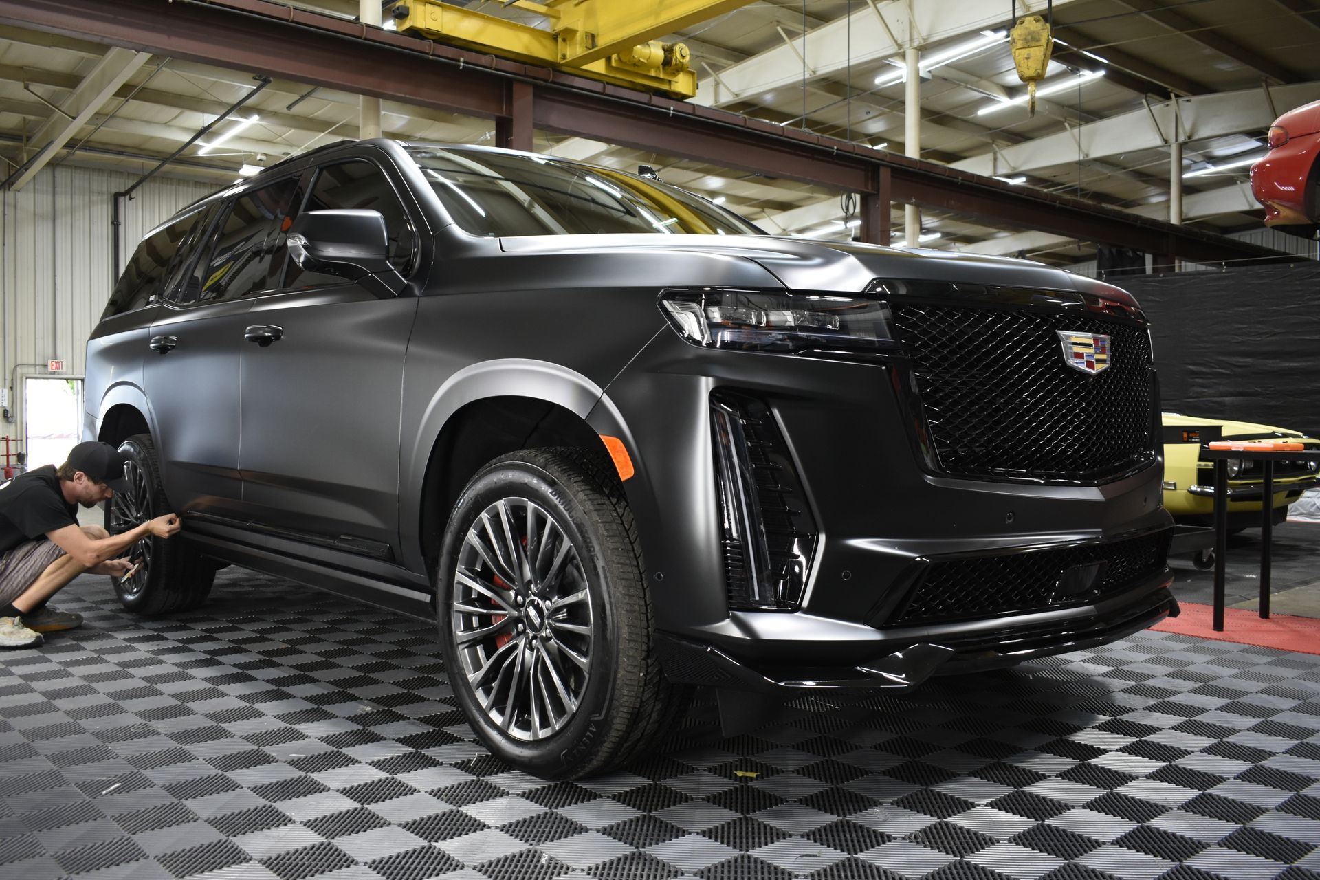 A matte black Cadillac Escalade SUV in a shop, with a person working on its wheels.