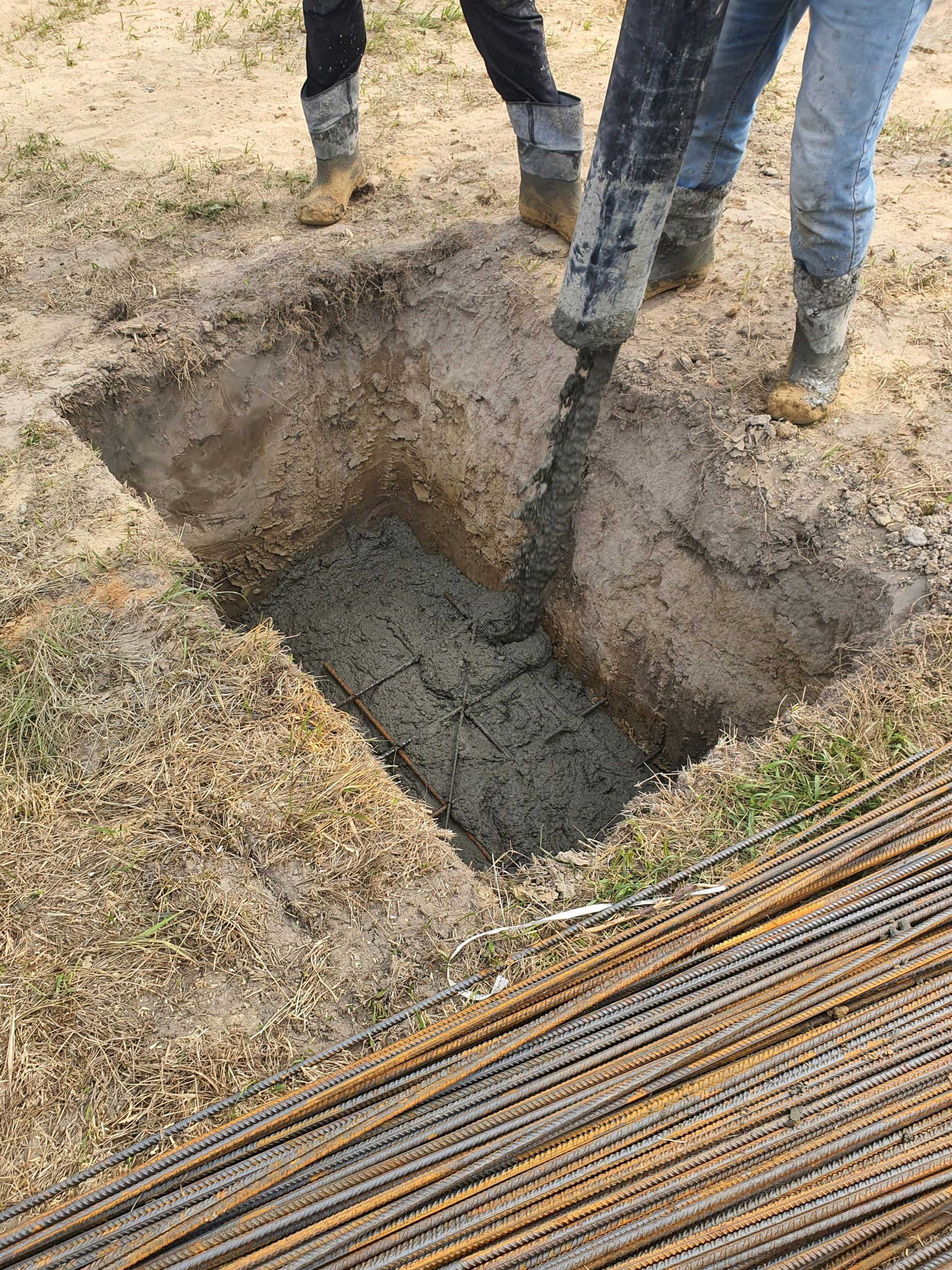 Concrete being poured from a hose into a square excavated hole in the ground, with metal rebar lying in the foreground.
