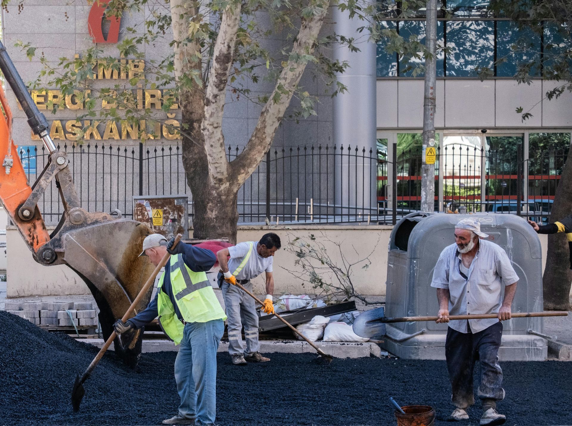 Three workers use shovels to spread fresh asphalt on a road near a building, with an excavator bucket visible nearby.