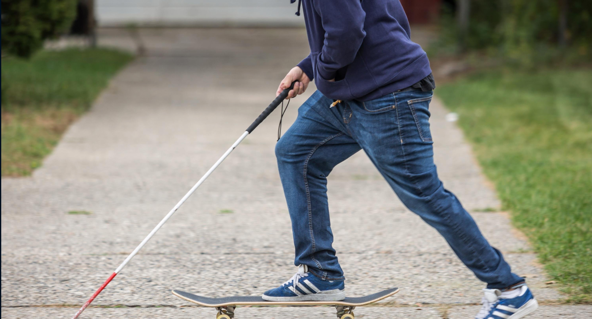 A blind skateboarder uses their cane to feel for obstacles and cracks in a sidewalk.