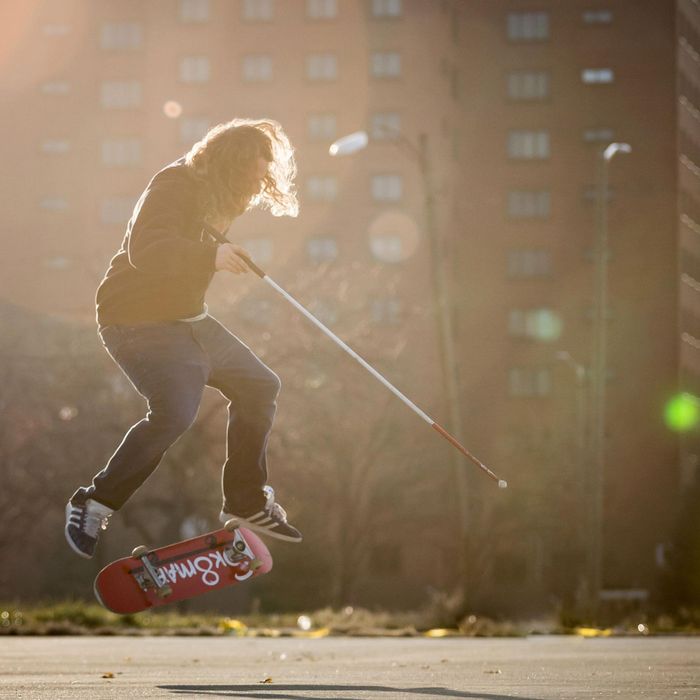 Dan Mancina is captured midair doing a flip trick on his skateboard, cane in hand.