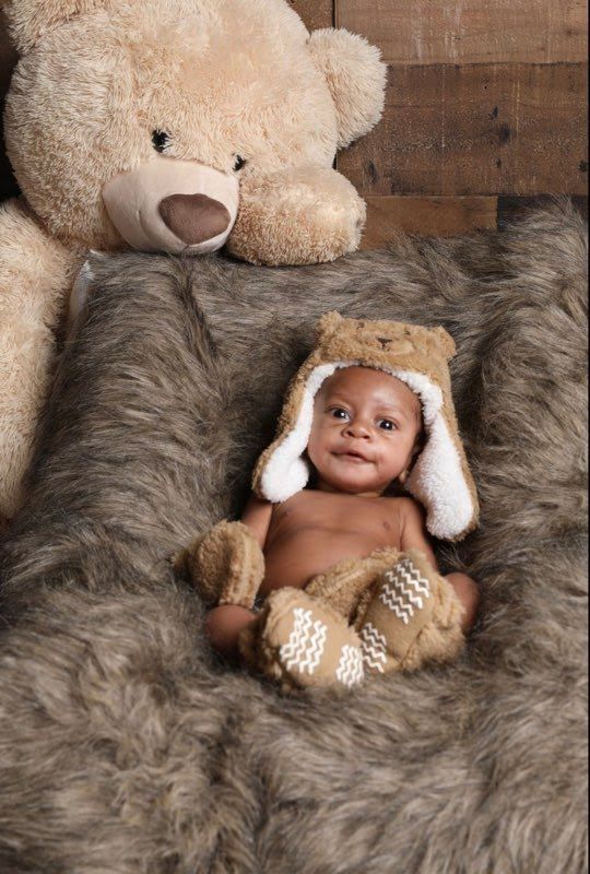 Baby wearing bear hat and mittens, sitting on fluffy brown rug, with a large teddy bear.