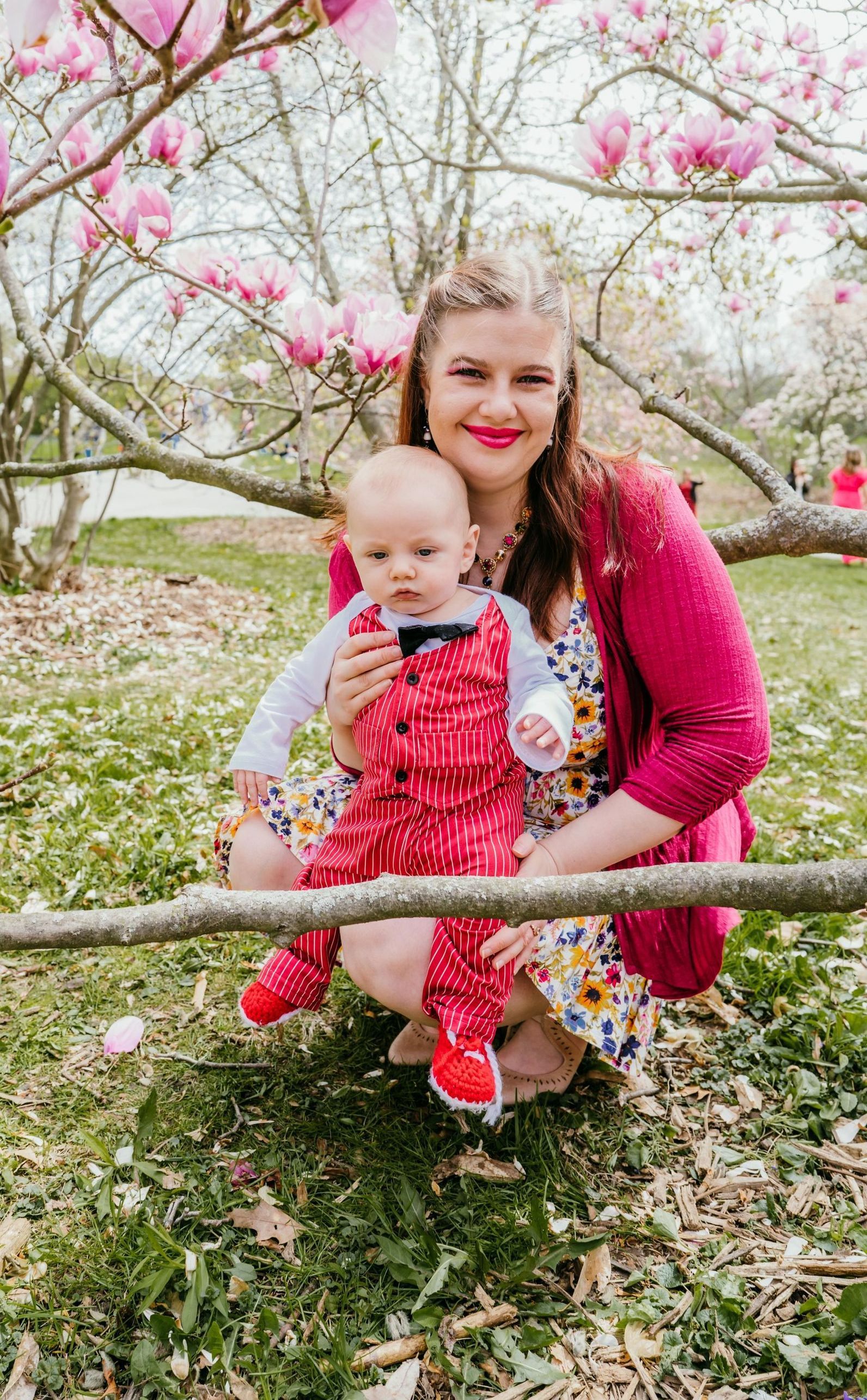 Woman in pink sweater, holding baby in red polka-dot overalls under blossoming tree.