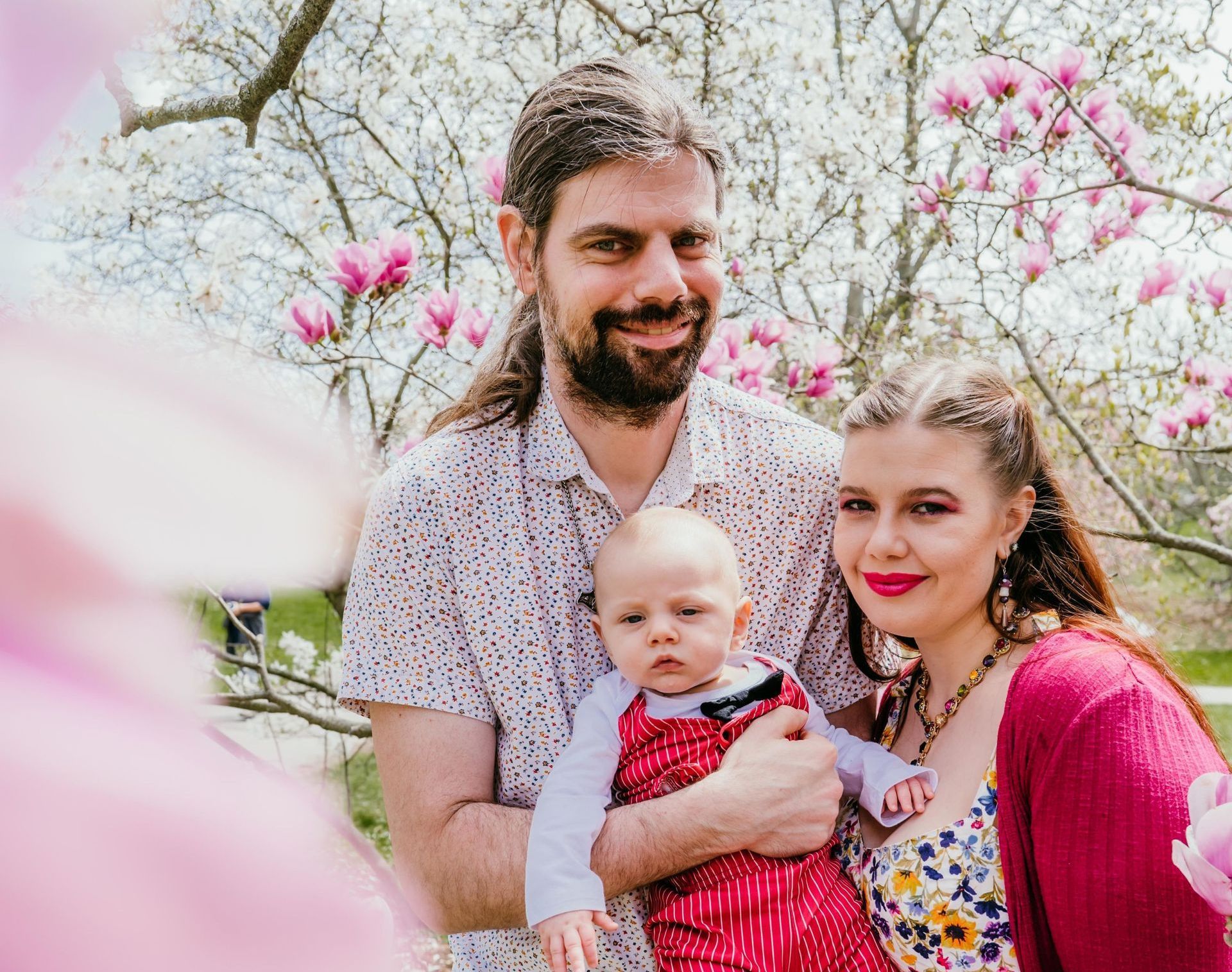Family of three smiling outdoors, baby in arms, flowering tree backdrop.