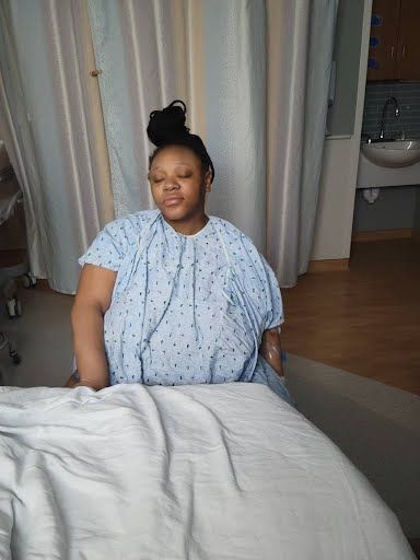Woman in hospital gown sitting on a bed, eyes closed. Hospital room setting.
