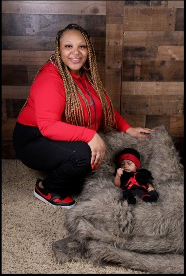 Woman in red outfit squats next to baby in a matching outfit on a gray fur rug. Wood backdrop.