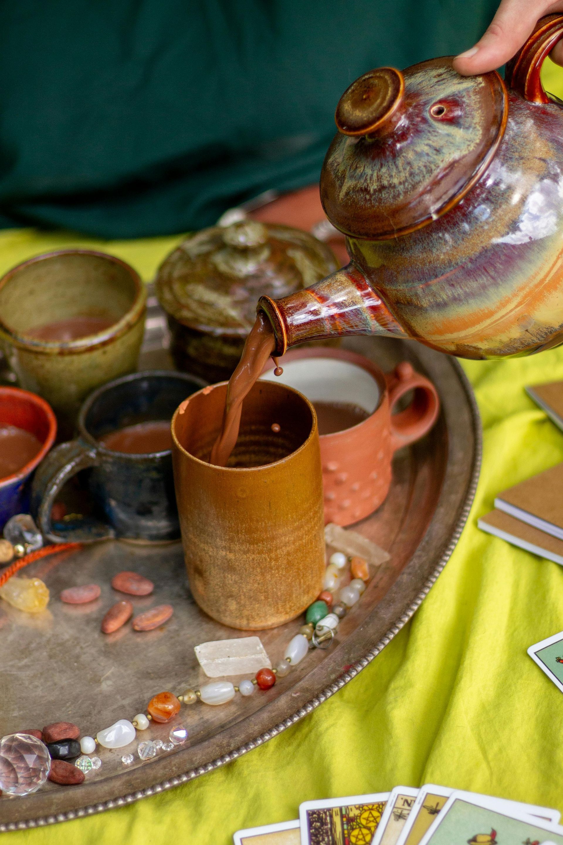 Person pouring chocolate drink from a ceramic pot into a cup on a tray with other mugs, beans, and jewelry on a yellow cloth.