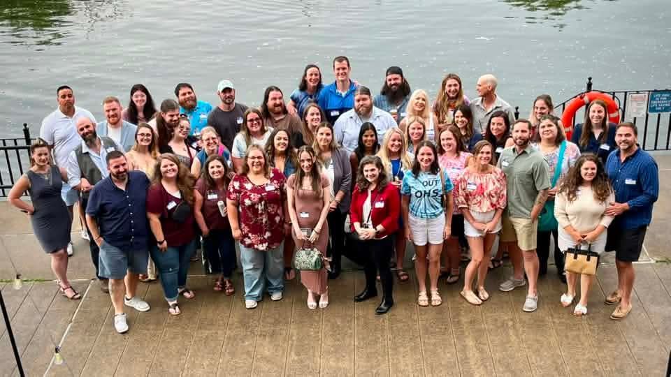 Large group posing for a photo on steps by a lake, with one person highlighted by a red circle.
