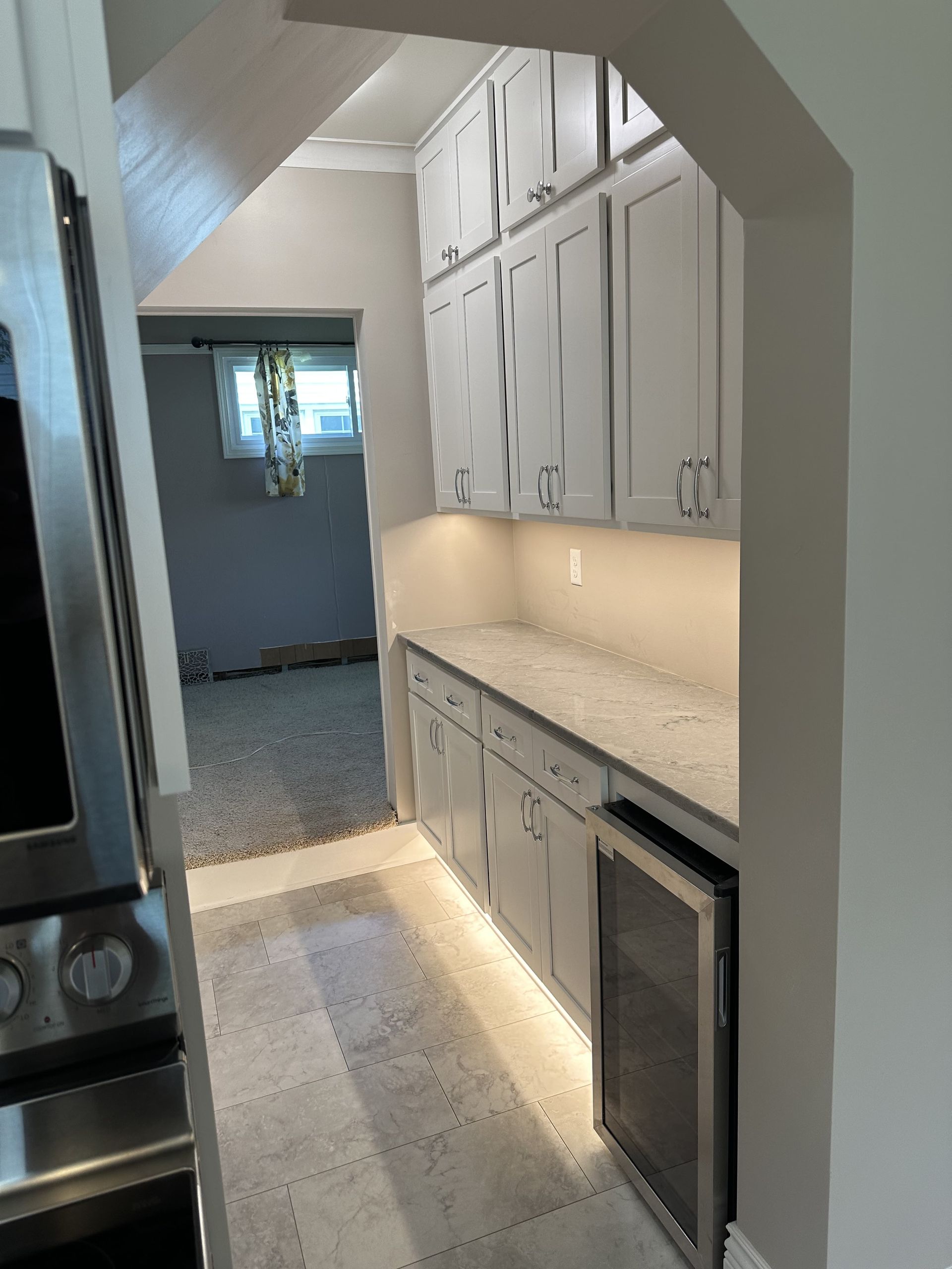 A narrow kitchen pantry with gray cabinets, countertop, and wine fridge. Soft lighting illuminates the space.