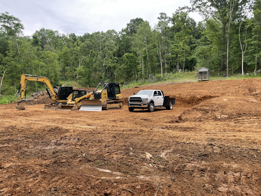 Construction site: excavator, pickup truck, and portable toilet on a dirt clearing, with trees in the background.
