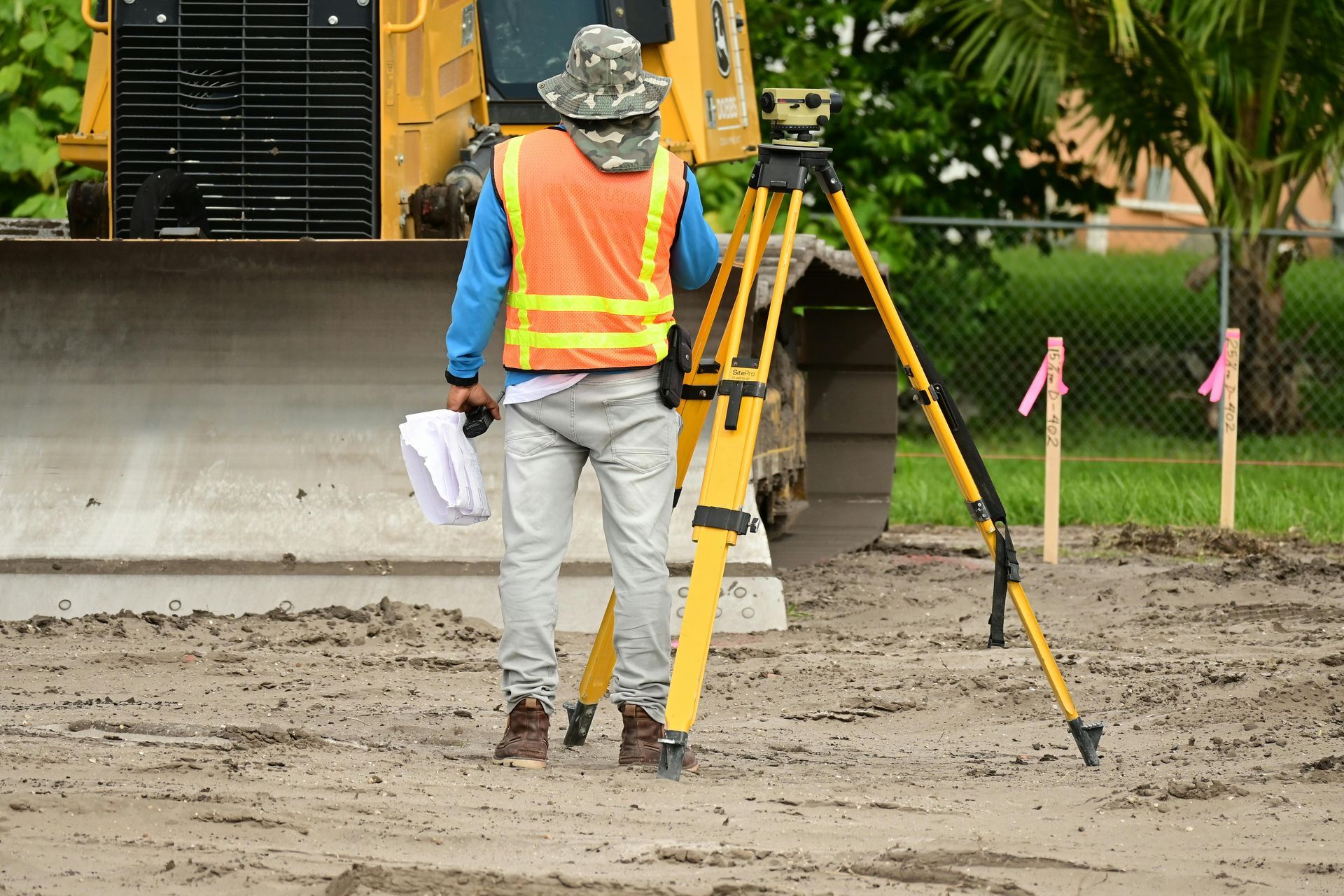Surveyor in an orange vest using a tripod and surveying equipment at a construction site.