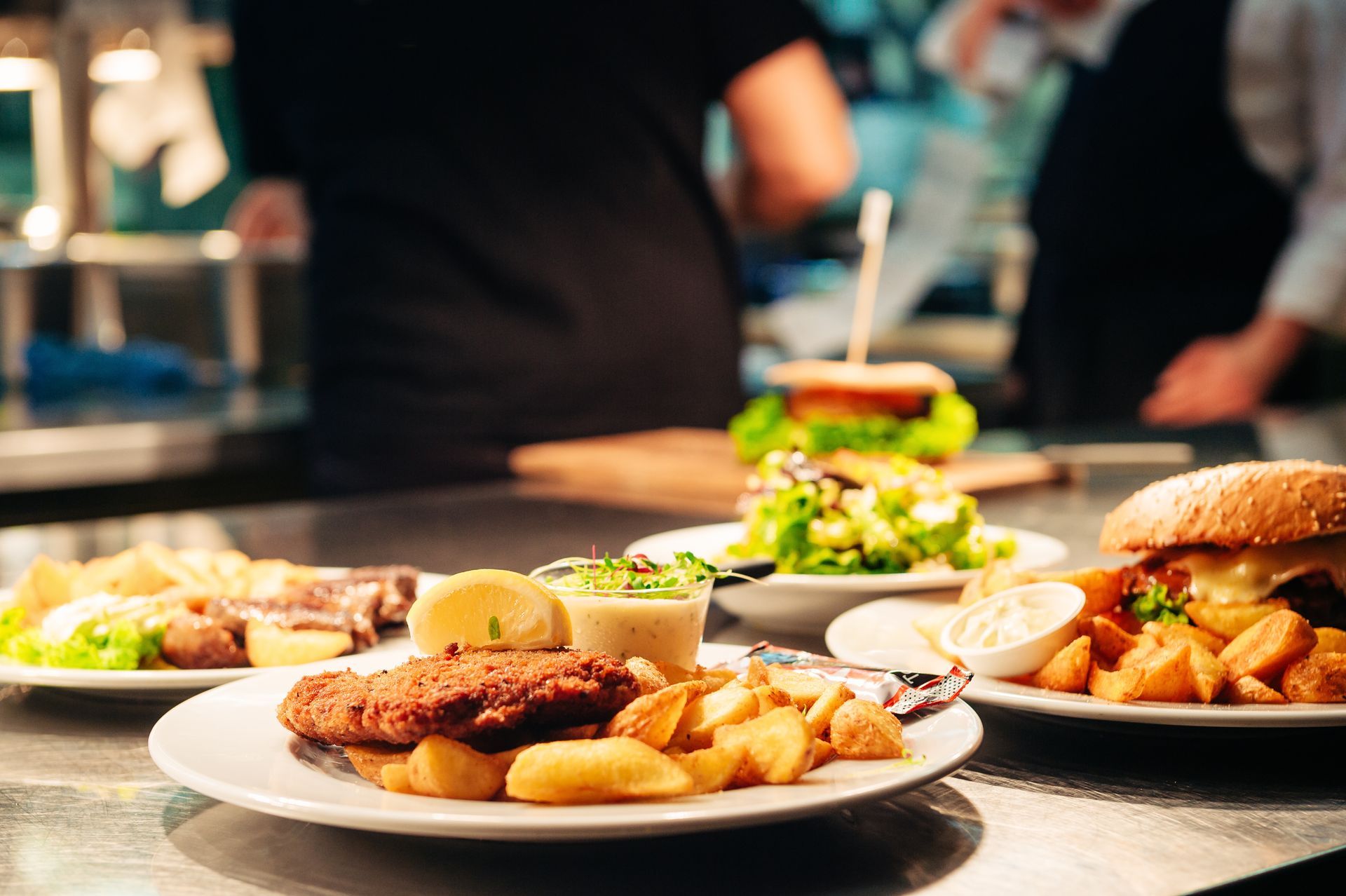 Plates of food on a counter in a restaurant kitchen: fried food, burgers, and salad. Chefs are blurred in the background.