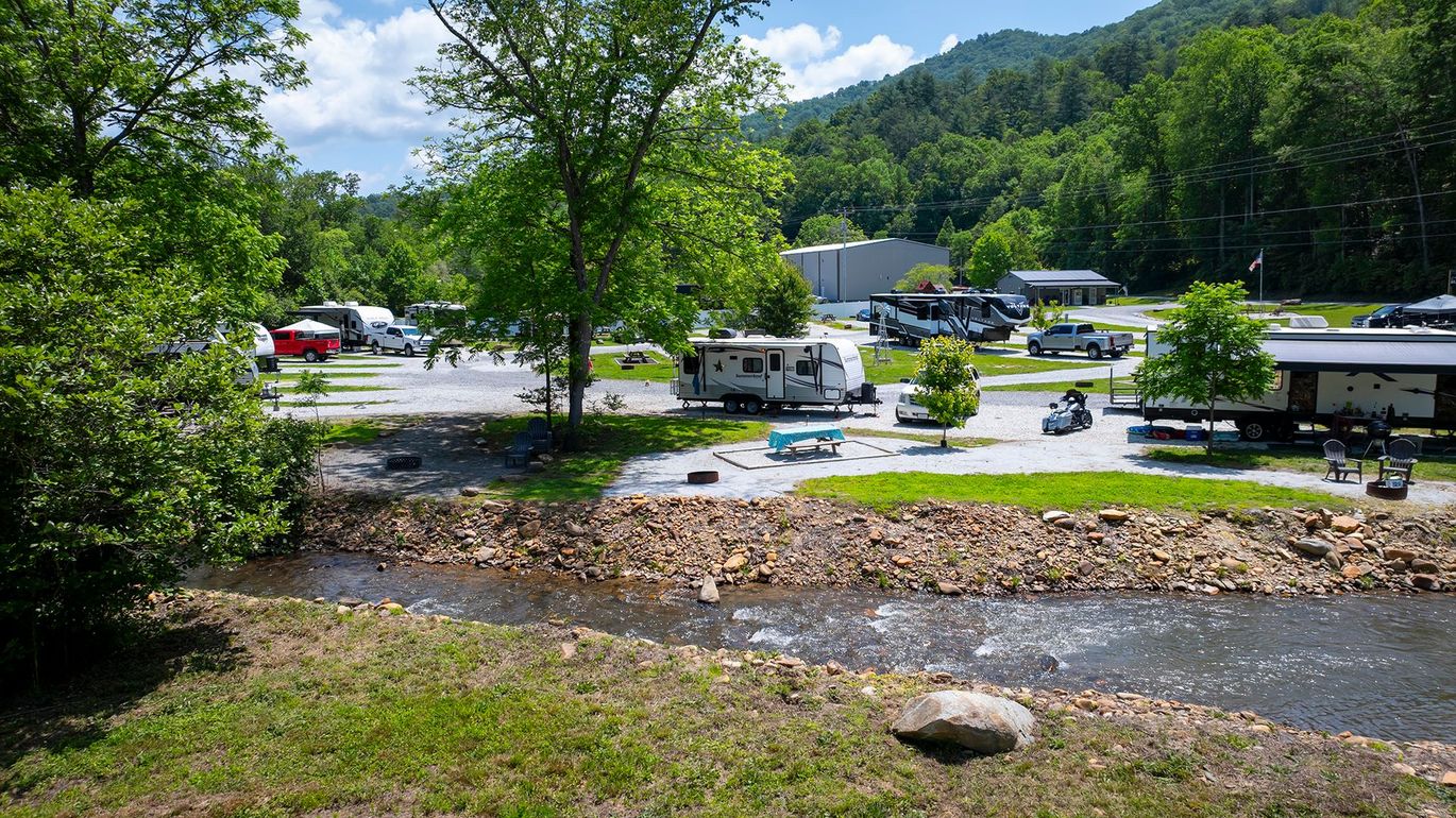 Campground scene with RVs parked near a flowing river, surrounded by trees and mountains on a sunny day.