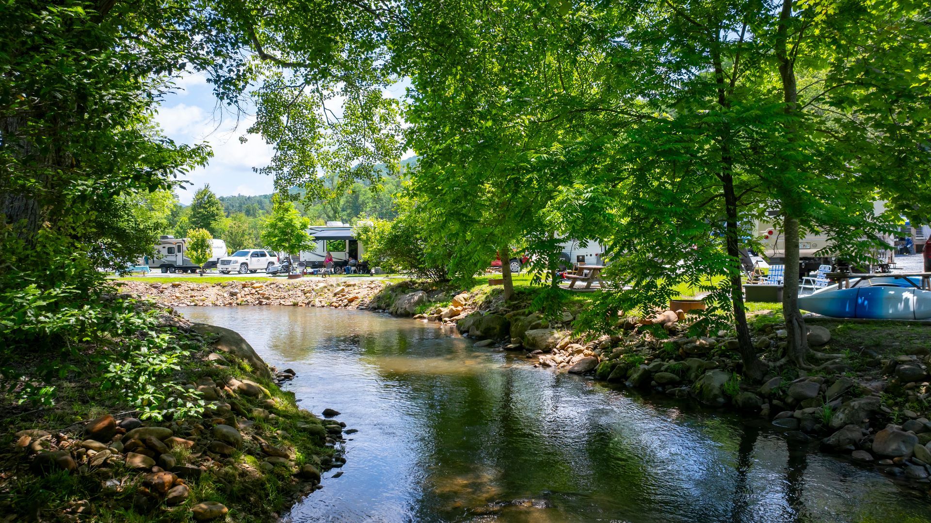 A tree-lined creek flows through a campground on a sunny day; RVs and a gazebo are visible in the distance.