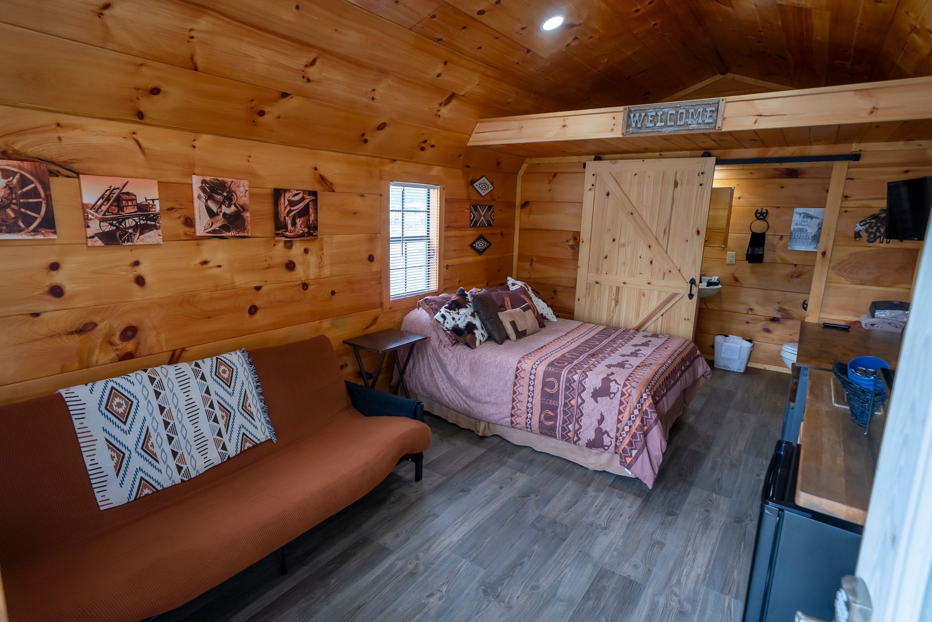 Cozy cabin interior with a bed, futon, artwork, and a sliding barn door to a bathroom. Wooden walls and floors are prominent.