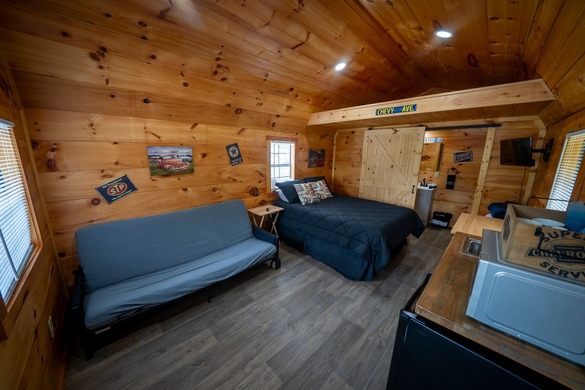 Cozy cabin interior with wooden walls and floors, featuring a futon, bed, and small appliances, including a mini-fridge.
