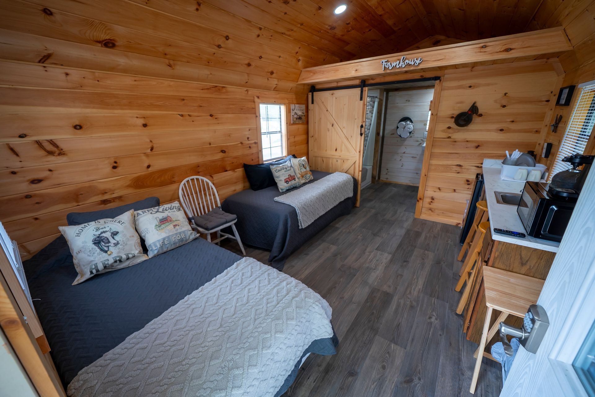 A small cabin bedroom with two beds, light wood walls, a sliding door to the bathroom, and a small table with a coffee maker.