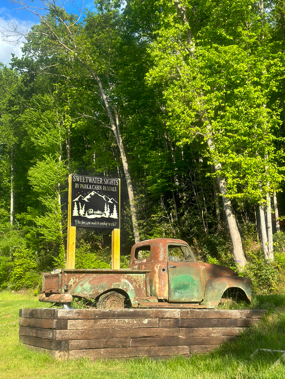 An old rusty truck is parked in front of a sign in the woods.