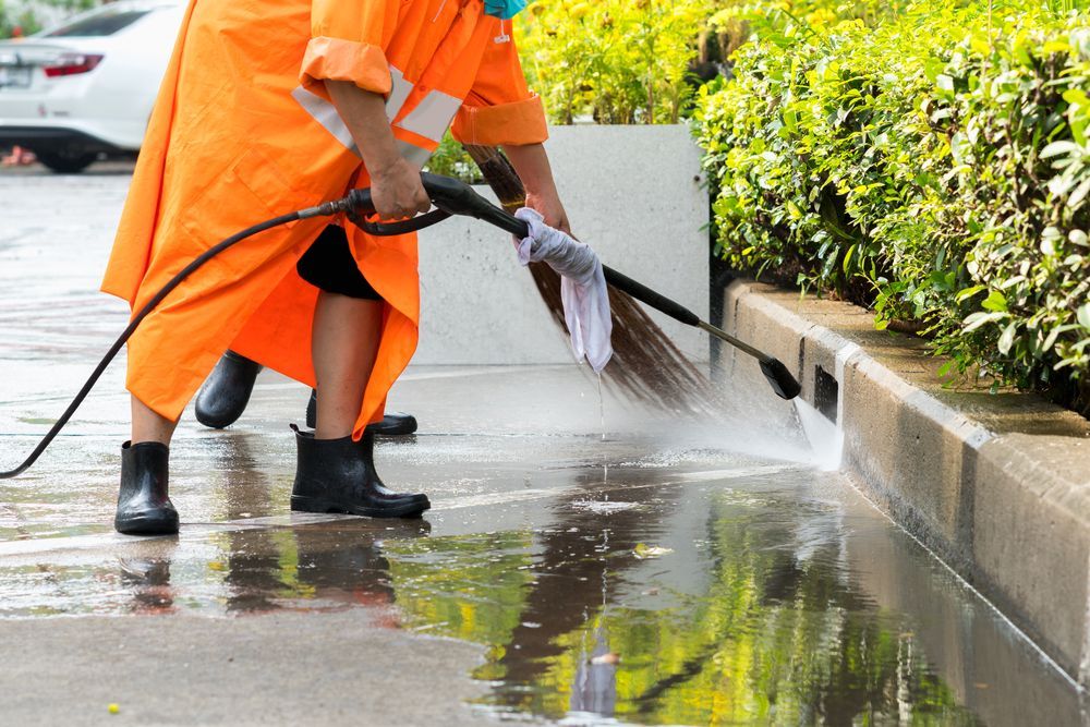 A man in an orange raincoat is cleaning the sidewalk with a high pressure washer.
