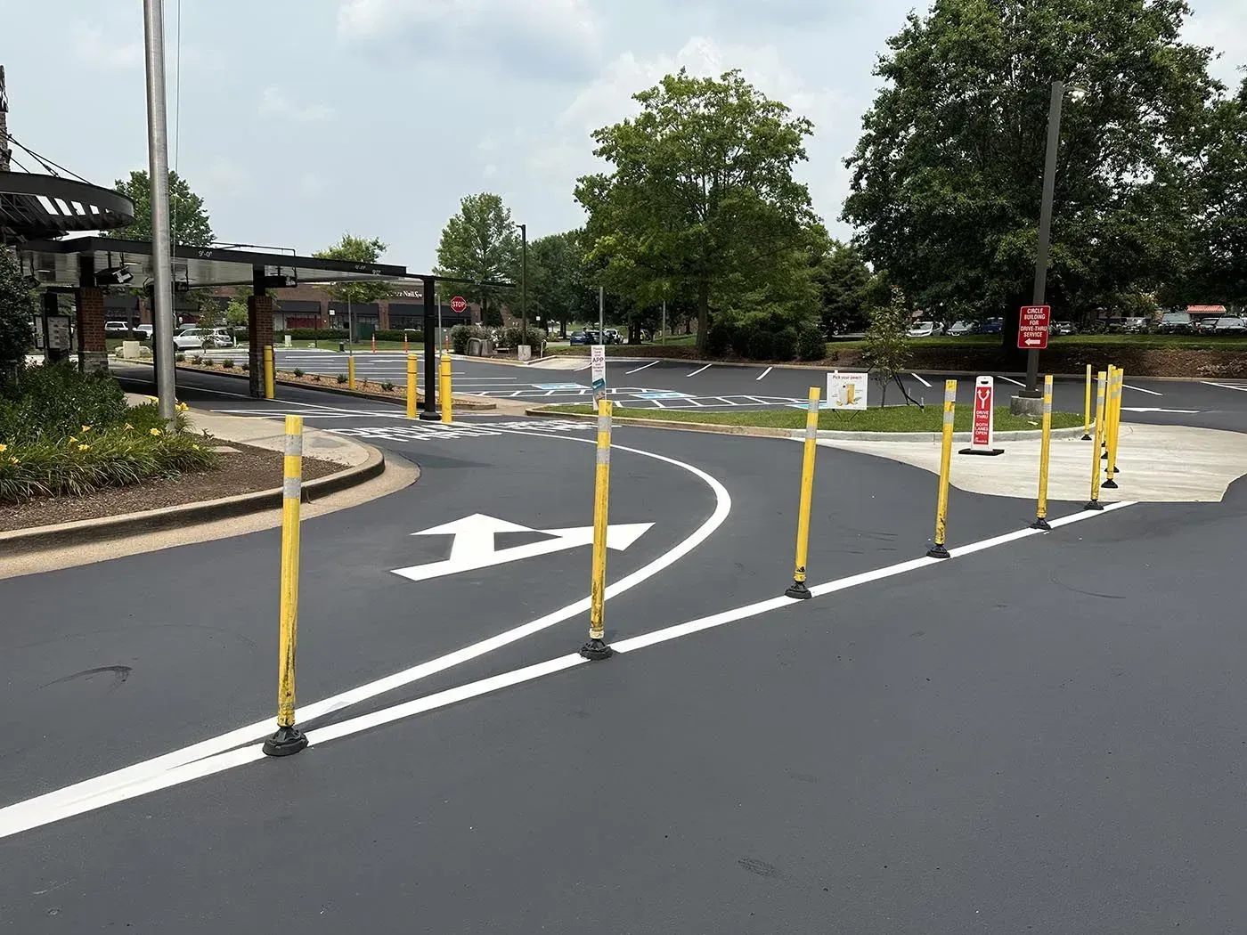 Drive-thru entrance at a restaurant, marked with yellow bollards and white arrows on fresh asphalt.