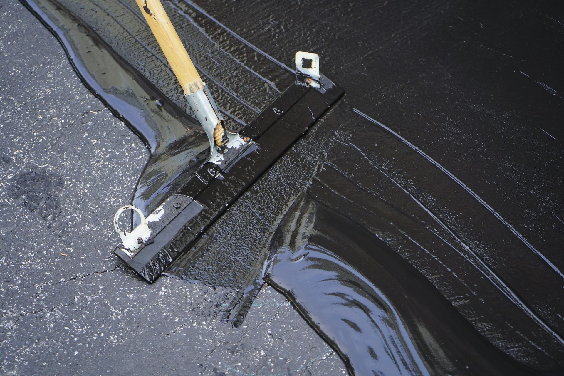 Worker spreading liquid asphalt sealant on pavement with a long-handled squeegee.