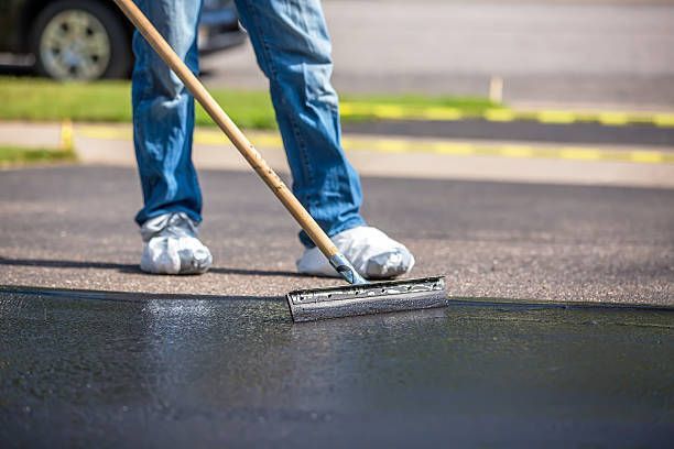 Person in jeans applying black sealant to a driveway with a squeegee. Sunlight, asphalt, and car are visible.