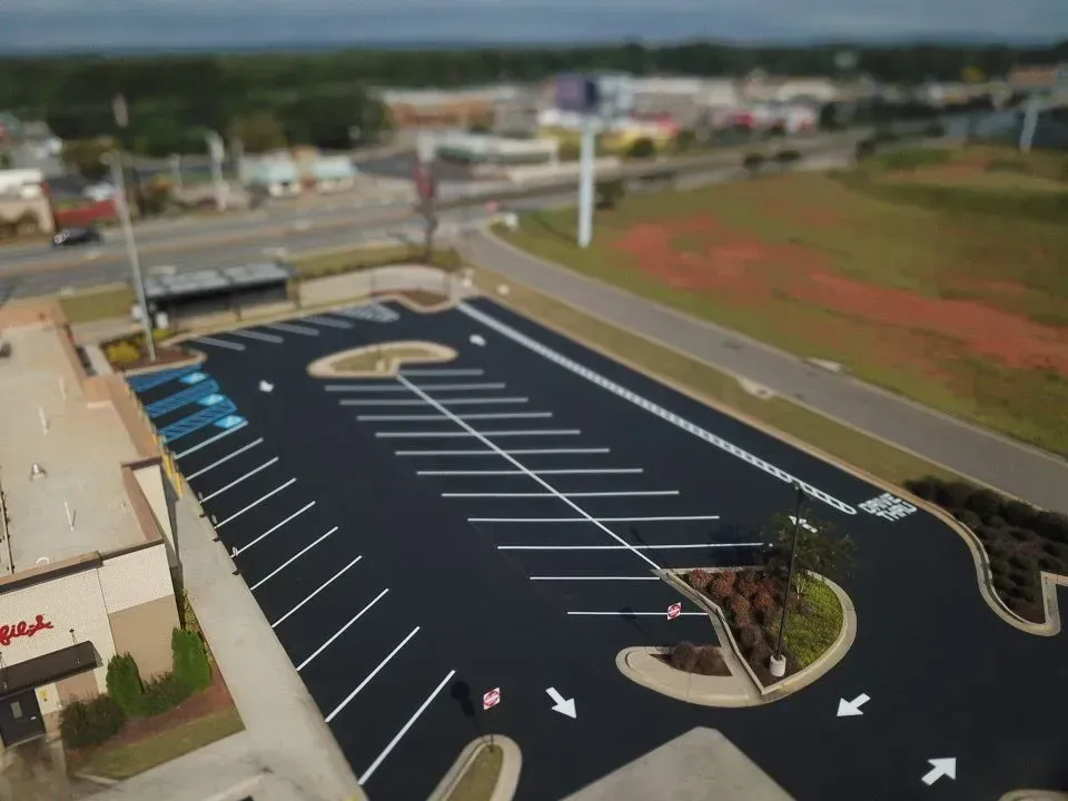 Aerial view of an empty black asphalt parking lot with white parking space lines and landscaping.