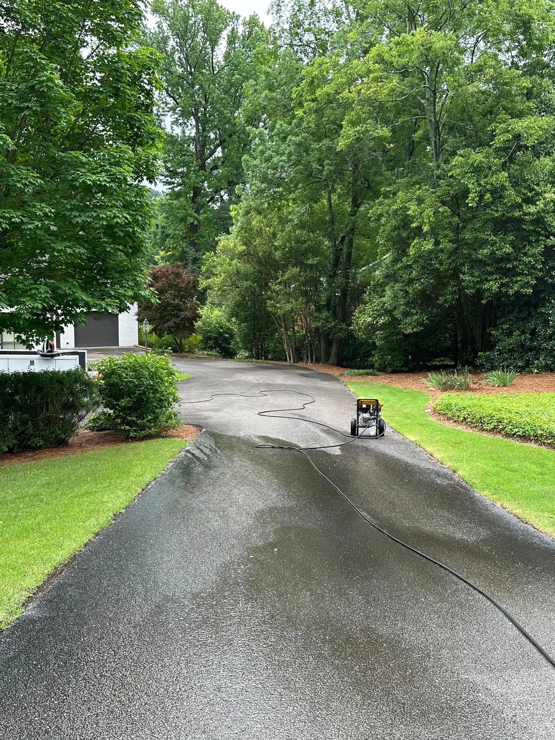 A person using a pressure washer to clean a wet, dark driveway, surrounded by green trees and grass.
