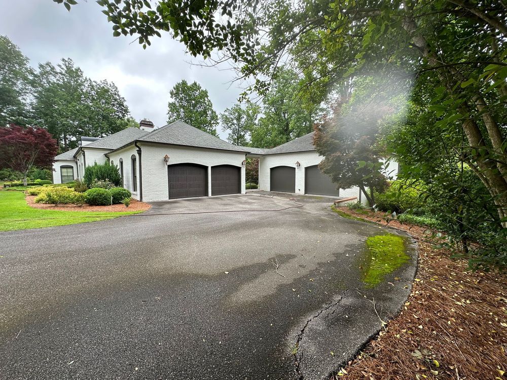 A white brick house with a three-car garage, brown doors, and a dark asphalt driveway, trees on the right.